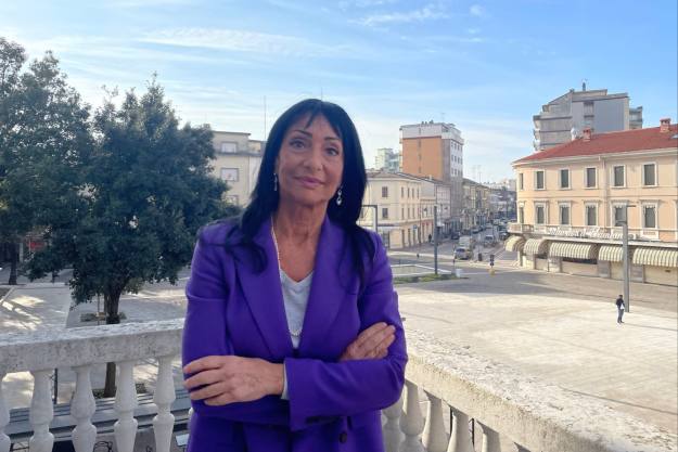 Anna Maria Cisint, wearing a purple jacket, stands with her arms folded, on a balcony with a view of Monfalcone in the background