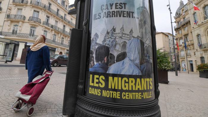 A woman walks by a poster reading "They are coming, Migrants in our town centre" in a street of Beziers.