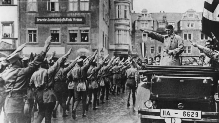 Nazi leader Adolf Hitler (1889 - 1945, in car, right) takes the salute as Sturmabteilung (SA) paramilitaries march past in the market square in Weimar, Germany, 13th November 1930. On the far right is Hitler's personal adjutant, Rudolf Hess (1894 - 1987). (Photo by Hulton Archive/Getty Images)