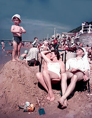 A couple relaxing on deck chairs as they enjoy the summer sun on the beach at Bournemouth as a small child stands on top of a large pile of sand (1952)