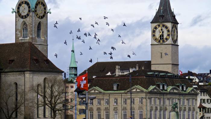 A Swiss national flag flies between the spires of Fraumeunster church and St Peter's Church in Zurich, Switzerland
