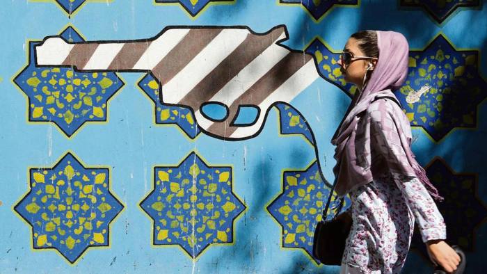 Iranian woman walks past a mural with a gun made out of an interpretation of the US flag