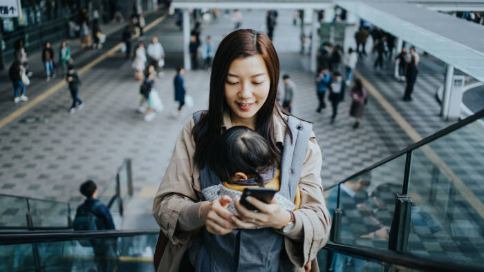 Young Asian mother with little daughter using smartphone while riding on escalator in downtown city, with busy commuters in the background