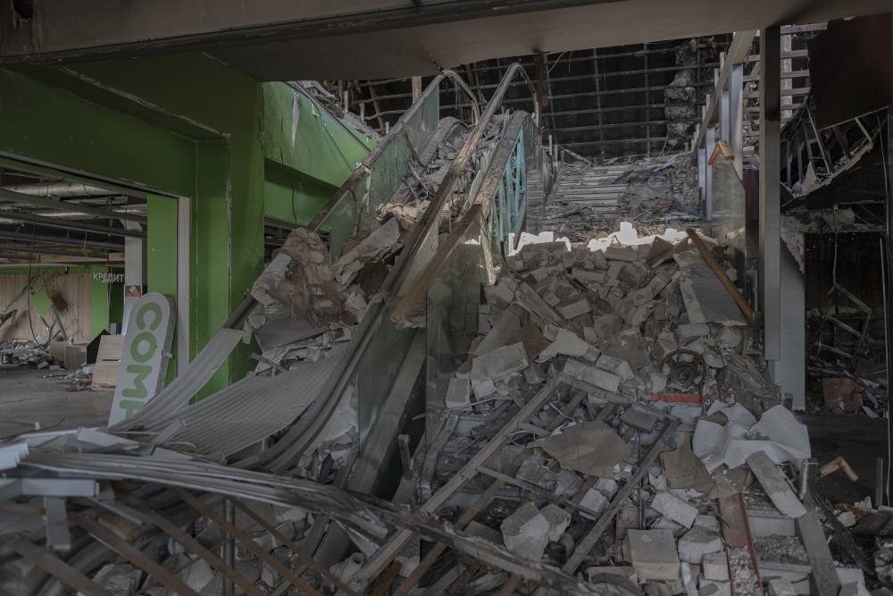 An image of the inside of the destroyed Giraffe mall. Rubble is piled up next to a destroyed staircase and escalator. Parts of walls and ceilings are missing.