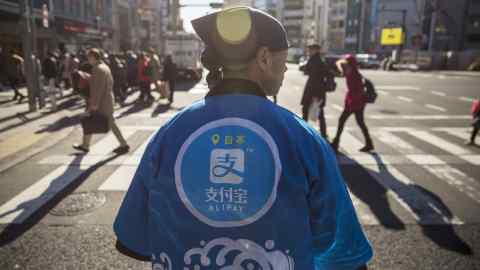 A staff member wearing a uniform featuring the logo for Ant Financial Services Group's Alipay, an affiliate of Alibaba Group Holding Ltd., stands during a campaign event in Tokyo, Japan, on Saturday, Dec. 9, 2017. Ant Financial and its strategic partners outside China should be able to nearly double users of their payments systems in coming years, Ant's overseas operations president Douglas Feagin said on Nov. 14. Photographer: Shiho Fukada/Bloomberg