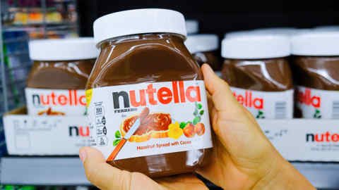 Shoppers hand holding a plastic jer of nutella brand hazelnuy spread with cocoa for sale  in a supermarket aisle