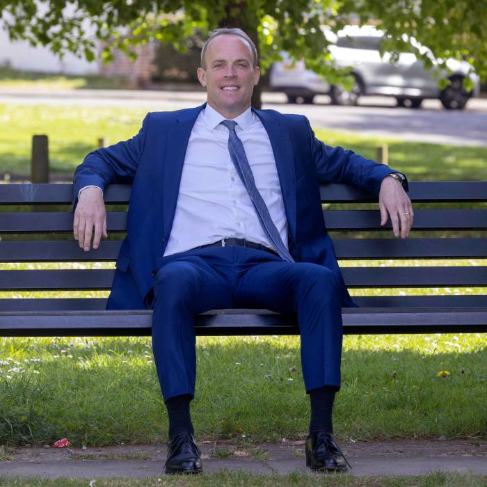 Dominic Raab, in suit and tie, sits smiling on a park bench on a sunny day