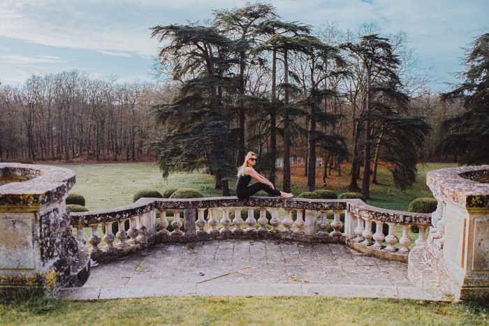 A woman sits on a stone terrace wall in front of acres of trees and lawns  