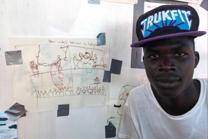 A rescued migrant called Adam poses next to his drawing of a torture scene at the Bani Walid compound in Libya