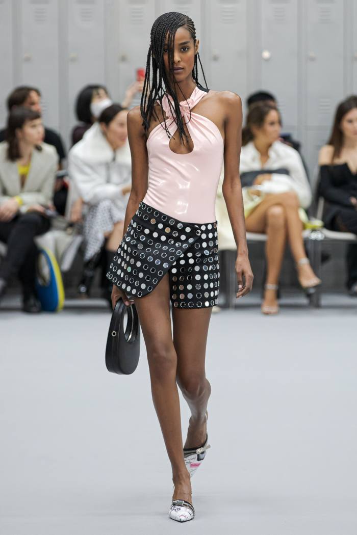 A model holds a handbag, wearing a pink top and a dark printed mini skirt