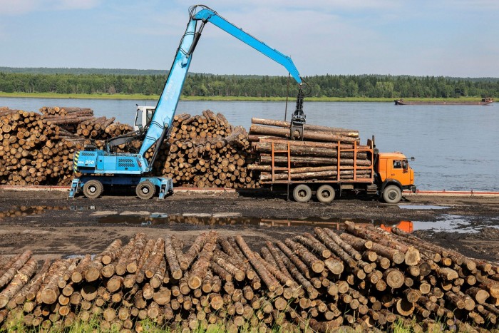 A truck-mounted log loader lifts logs from a barge docked on the banks of the Yenisei River