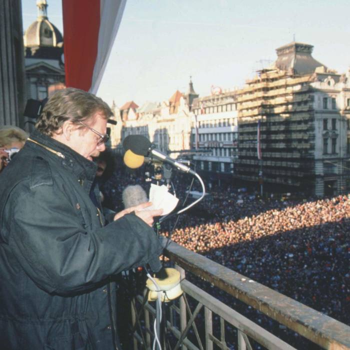 A man on a balcony speaks into a microphone, as crowds below listen