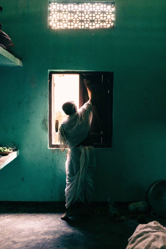 An interior shot of a darkened room with green walls, and man at the window reaching up to open the curtains