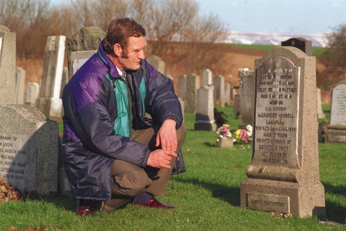 A man in a puffer jacket kneels by a tombstone