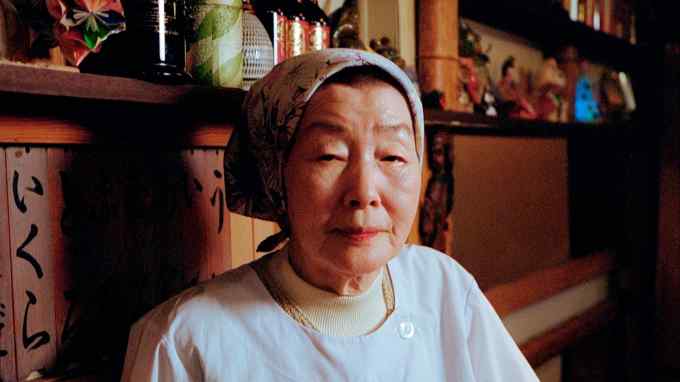 Sushi chef Sachiko Shimizu wearing a white apron and a headscarf in front of shelves filled with ornaments