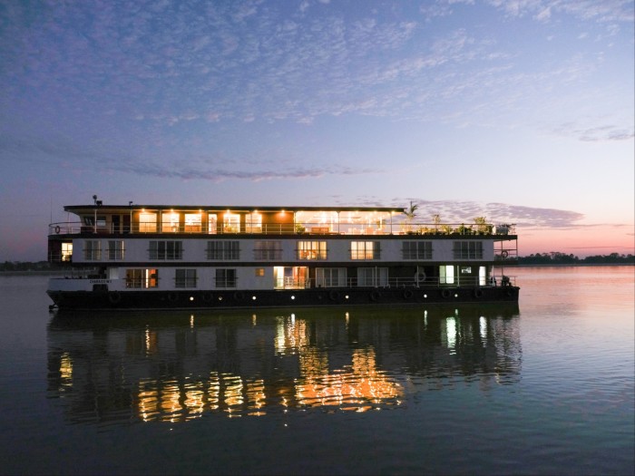 A river cruise boat at dusk, with cabins lights aglow and a pink sunset in the background 