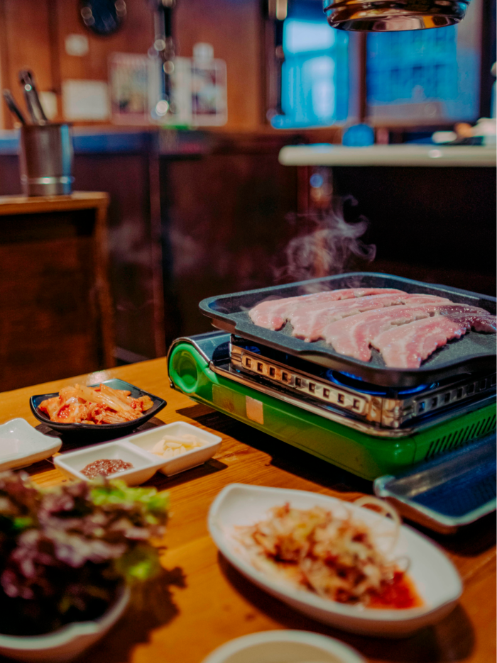 Pork on the grill at a table in Tomato Restaurant, surrounded by plates of kimchi and lettuce and dipping sauce