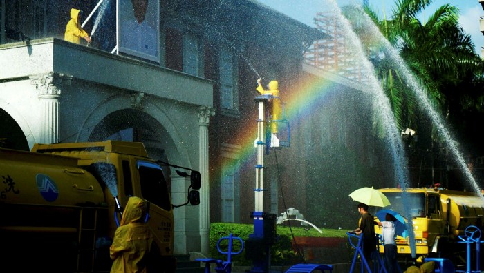 Water trucks are parked outside a building while people in yellow rain jackets hold hoses spraying water, creating a rainbow
