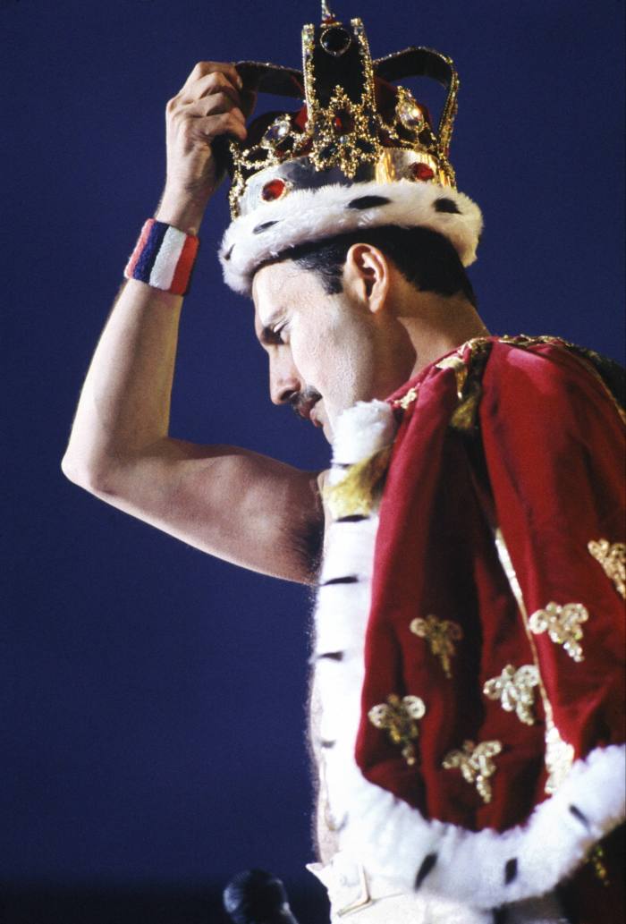 Freddie Mercury on stage wearing an ermine trimmed crown and red velvet cloak, also with ermine trim