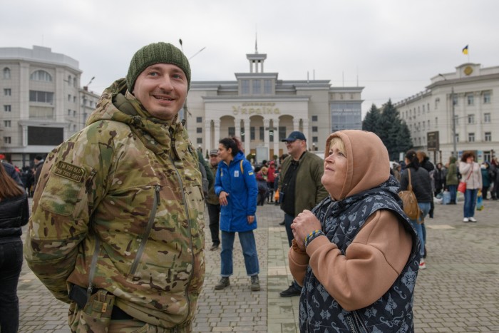 A woman stands with her hands clasped, looking up at a smiling man wearing military fatigues