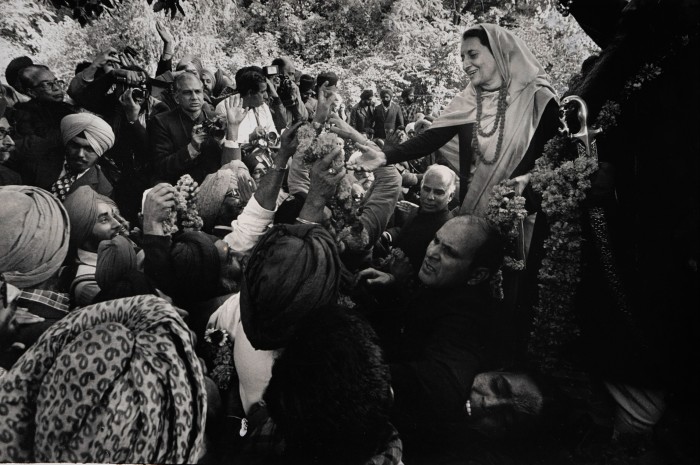 A photograph taken in India shows a crowd of men carrying garlands of flowers and cameras clustering around a woman who smiles and reaches out to them