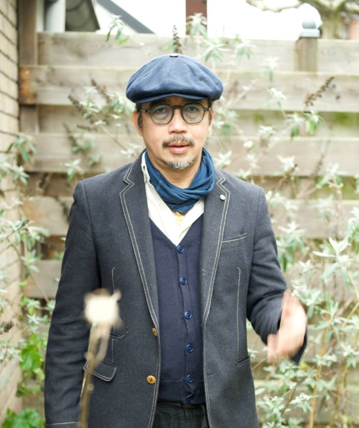 A man with a goatee wearing a flat cap stands in a patio setting and gestures towards the camera