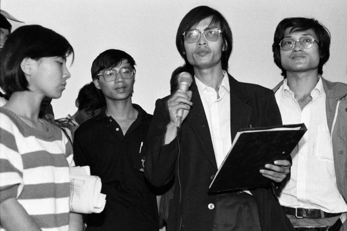 Feng Congde, a student leader, talks into a microphone, surrounded by three other leaders, including Li Lu. The group is announcing their intention to stay in Tiananmen Square throughout June 1989