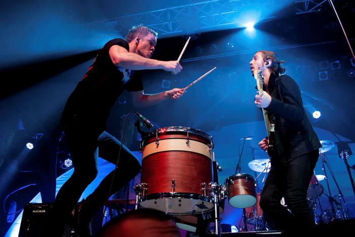 A man lifts his drumsticks high above a drum while another plays guitar