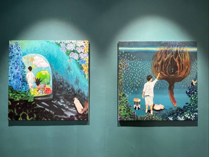 An installation view of two small, square paintings both featuring surreal garden scenes with a young boy as protagonist