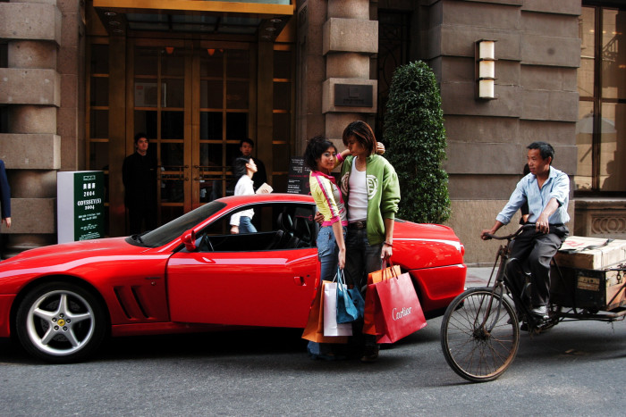 A young couple with boutique bags stand by their Ferrari while an older, poorer man, a courier, rides past on his bike loaded with boxes