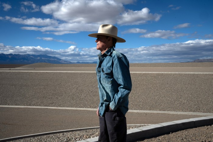 A man in a blue shirt and stetson stands in a desert landscape with a blue sky behind him.