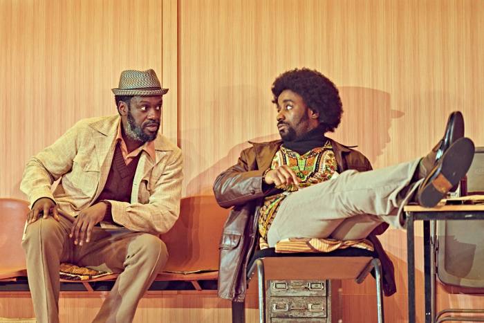 Two black men wearing 1970s clothing sit and talk animatedly in a bare-looking room