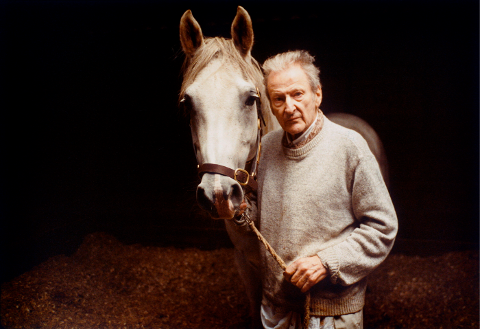 Um homem grisalho está segurando as rédeas de um cavalo.  Ambos encaram a câmera