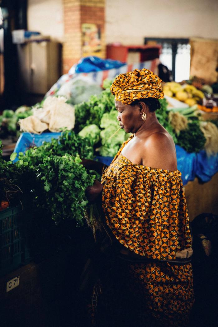 A woman in a richly coloured dress and matching headgear peruses vegetables at a market stall