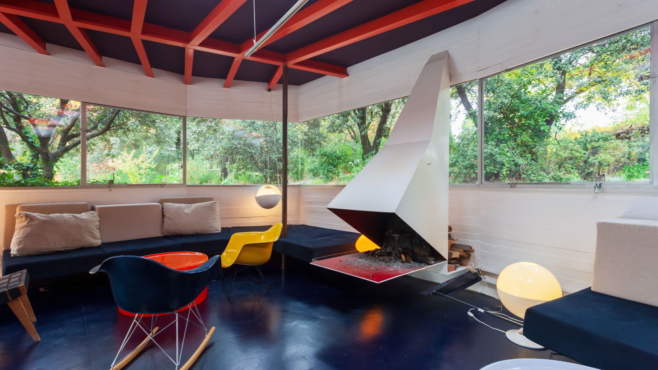 orange, red and yellow colour accents in a mostly blue contemporary sitting room with wraparound windows with greenery showing through