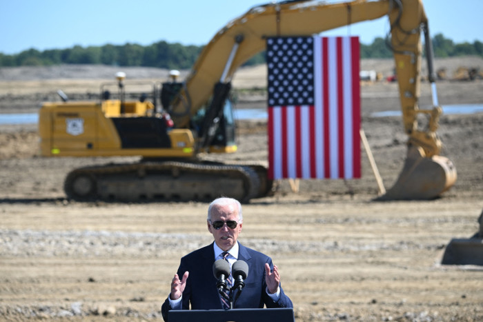 Joe Biden stands at a podium in front of an empty building site where a digger displaying the US flag prepares to break ground
