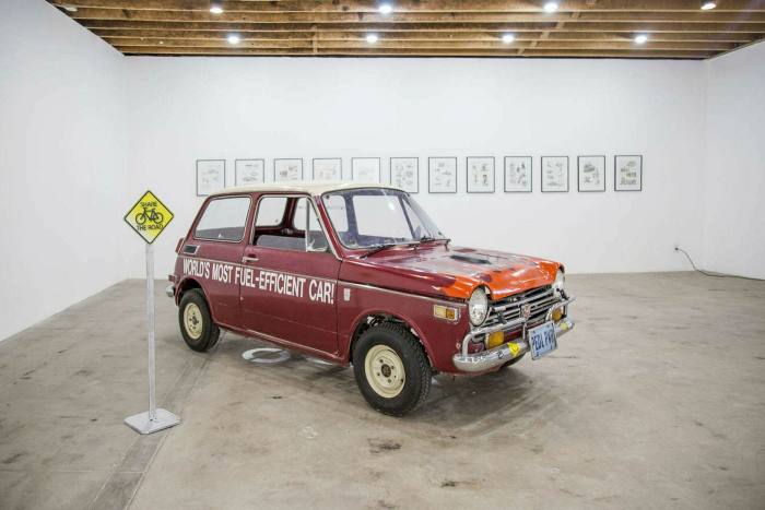 An old car in an art gallery. Text on the car’s side says: world’s most fuel-efficient car!