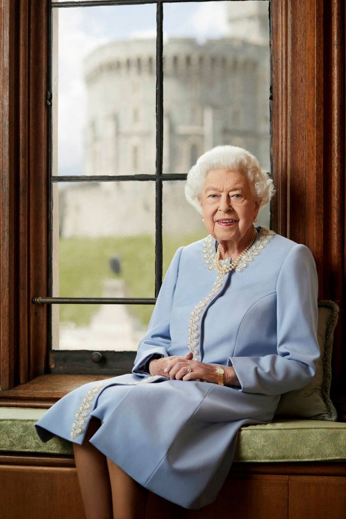 La reine, vêtue d'un habit bleu, est assise sur un siège de fenêtre.  Elle sourit et a les mains jointes sur ses genoux.  Par la fenêtre, nous pouvons voir de l'herbe et une tour ronde en pierre