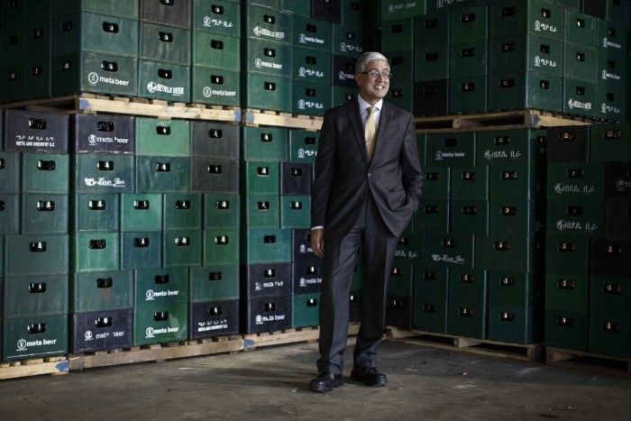 Ivan Menezes in suit and tie, standing smiling in front of stacks of crates of beer