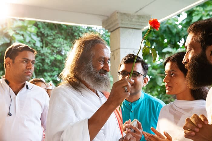 Shankar, in his white robes, holding up a long-stemmed red rose. He is with a small entourage at his ashram