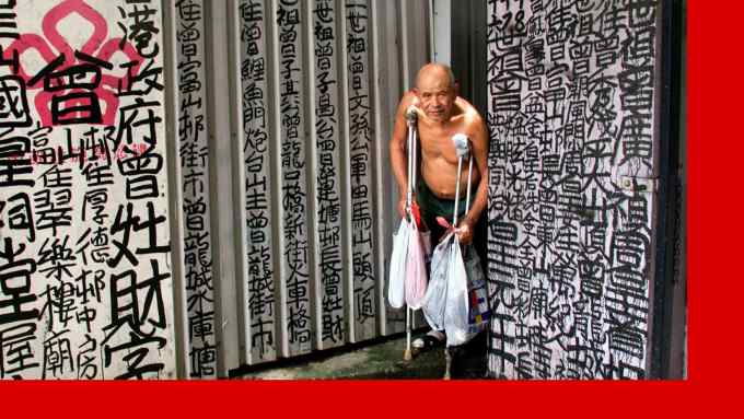 A shirtless older man stands on crutches and holds plastic bags, surrounded by walls covered in Chinese calligraphy