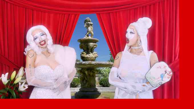Two individuals are stood in front of a red curtain with a statue behind it. Both wear white wigs and ballgowns and have exaggerated make-up or face paint.