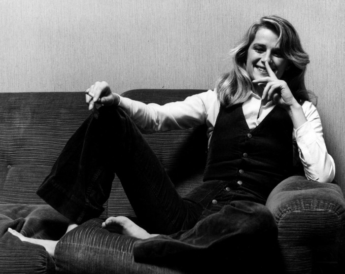 The actress Charlotte Rampling sits back on a sofa, barefoot. She wears a waistcoat, white shirt and trousers