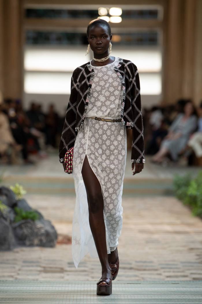 A model walks the runway wearing a white dress with a thigh slit and a black and gold cardigan