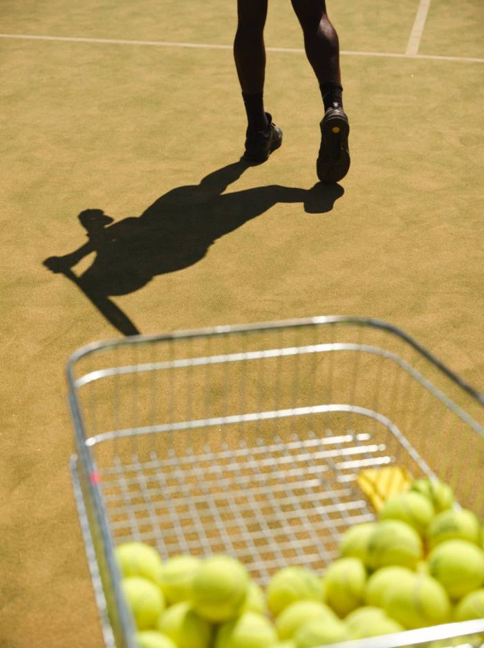 Een mand vol tennisballen achter de benen van William Ikome en zijn schaduw op een tennisbaan