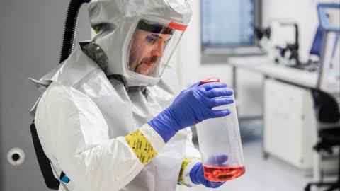 Un técnico de laboratorio con un traje de protección de cuerpo completo manipula un vial que contiene medios de crecimiento para la producción de virus