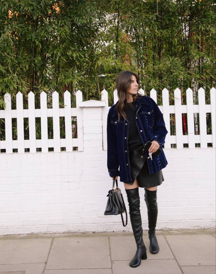 A woman in a short skirt, blue jacket and long boots stands in front of a white picket fence 