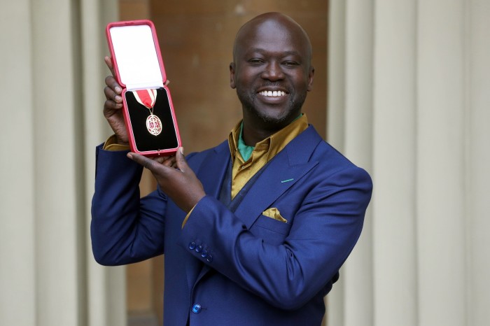 Sir David Adjaye smiling, wearing a blue suit, and holding up his knight’s insignia