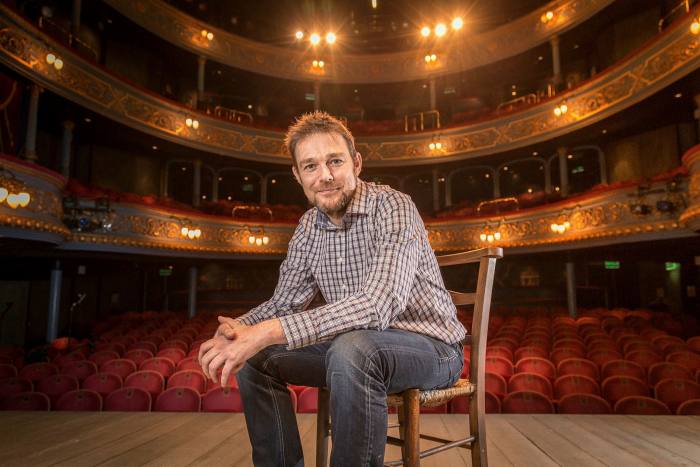 A man in casual clothes sitting on a chair on a stage inside a theatre