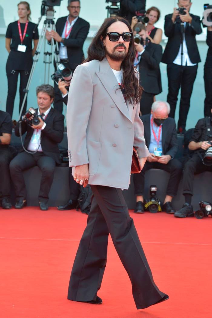 Alessandro Michele, who has long dark hair and a beard, walks on a red carpet with photographers behind him